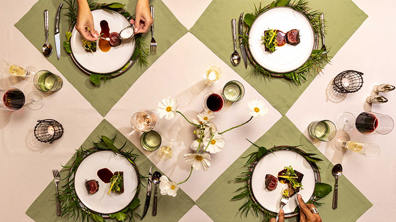 Overhead view of an elegantly set dining table with four plated dishes on sage green linens, surrounded by fresh greenery, white flowers, wine glasses, water glasses, silverware, and candlelight. Two diners’ hands are visible interacting with their plates.