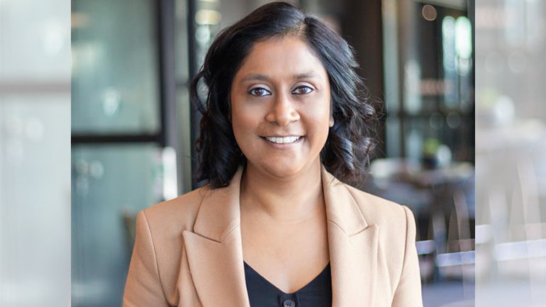Professional portrait of a woman wearing a beige tailored blazer over a dark top, standing indoors in a modern hospitality setting with soft lighting, glass features, and tables visible in the background.