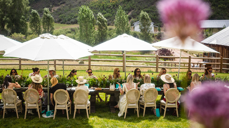 A long outdoor dining table set on a grassy lawn hosts a group of guests seated beneath large white umbrellas. The table is decorated with lush green floral arrangements, while blurred pink flowers frame the foreground. A wooden fence, tall trees, rustic buildings, and rolling green hills create a scenic countryside backdrop under bright daylight.