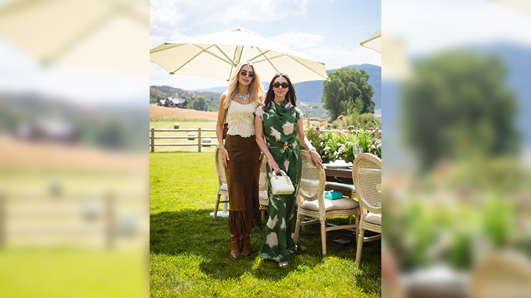 Two people wearing sunglasses pose beside a long outdoor dining table decorated with flowers on a green lawn. Large white umbrellas provide shade, while a wooden fence, rolling hills, trees, and distant mountains create a scenic countryside backdrop under a bright, partly cloudy sky. One person wears a cream lace top with a brown skirt and boots, while the other wears a green patterned dress and carries a small white handbag.