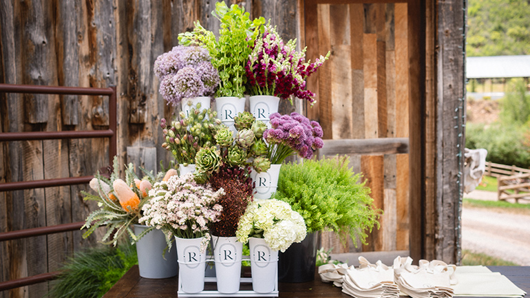 A rustic wooden table displays an assortment of freshly cut flowers and greenery arranged in white containers and metal buckets outside a weathered barn. The colorful blooms include lavender, pink, white, and deep magenta flowers alongside succulents and bright green foliage. Neatly folded canvas tote bags rest on one end of the table, while the barn's wooden siding and doorway create a charming countryside backdrop.