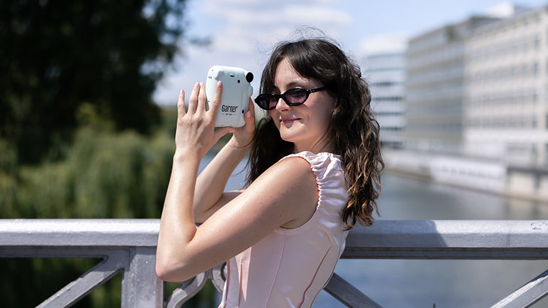 Person wearing dark sunglasses stands on a bridge overlooking a riverside cityscape, holding a light-blue Garner-branded instant camera. Trees, modern buildings and a waterway are visible in the background on a sunny day.