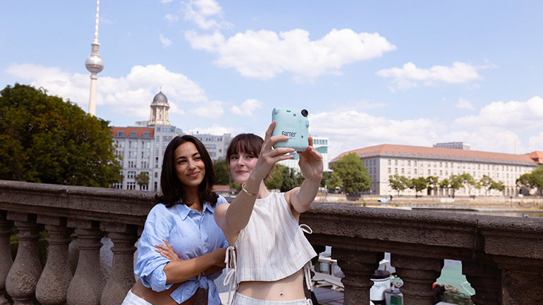 Two people take a photo with an instant camera on a riverside bridge in Berlin, with the TV Tower and city skyline in the background.