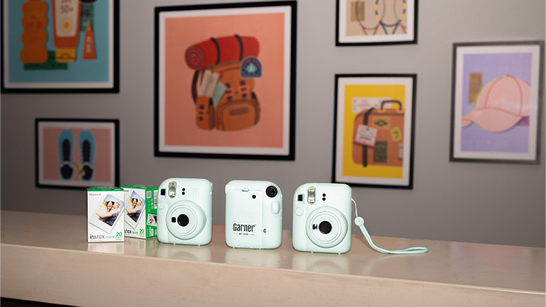 Three light-blue instant cameras and packs of instant film displayed on a reception counter in a modern hotel interior. Framed travel-themed artwork is visible on the wall in the background.