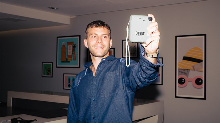 Person standing indoors holding a light-blue Garner-branded instant camera out in front of them. Framed artwork decorates the walls of the modern hotel interior in the background.