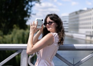 Person wearing dark sunglasses stands on a bridge overlooking a riverside cityscape, holding a light-blue Garner-branded instant camera. Trees, modern buildings and a waterway are visible in the background on a sunny day.