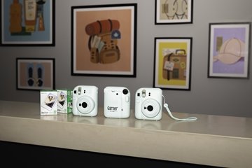 Three light-blue instant cameras and packs of instant film displayed on a reception counter in a modern hotel interior. Framed travel-themed artwork is visible on the wall in the background.