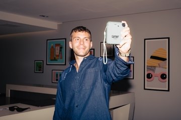 Person standing indoors holding a light-blue Garner-branded instant camera out in front of them. Framed artwork decorates the walls of the modern hotel interior in the background.