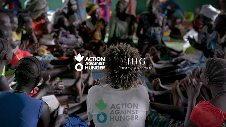 A group of people, including women and children, sit closely together on the floor inside a shelter, engaged in a communal activity. The photo is taken from behind a person wearing a white t-shirt with the logo and name of the humanitarian organisation "Action Against Hunger." In the center of the image are the logos of "Action Against Hunger" and "IHG Hotels & Resorts," indicating the partnership. The atmosphere suggests a humanitarian or community support setting.