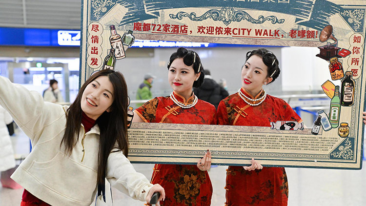 A woman takes a selfie with two performers in traditional Chinese dresses holding a decorative IHG photo frame at an event, likely in a public space such as a train station or airport.