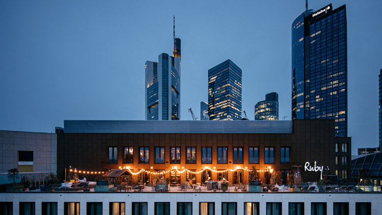 A nighttime photo shows the Ruby Hotel in Frankfurt, Germany, with its illuminated façade and outdoor seating area, set against a backdrop of the city’s modern skyline featuring several tall skyscrapers.